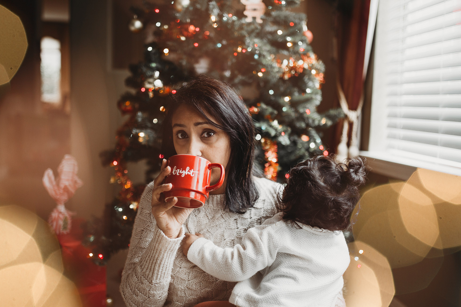 woman holding child and red coffee mug in front of christmas tree jyo bhamadipati